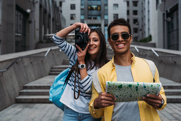 Woman taking photo on digital camera near bi-racial friend holding map