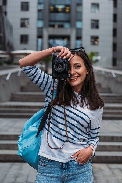 happy girl taking photo on digital camera and standing with hand in pocket outside 