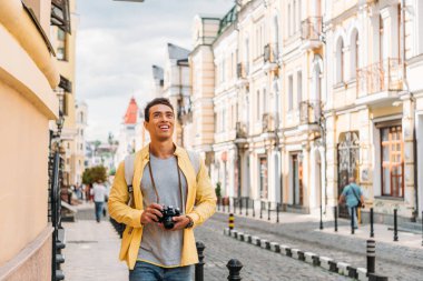 happy mixed race man smiling while holding digital camera near buildings  clipart