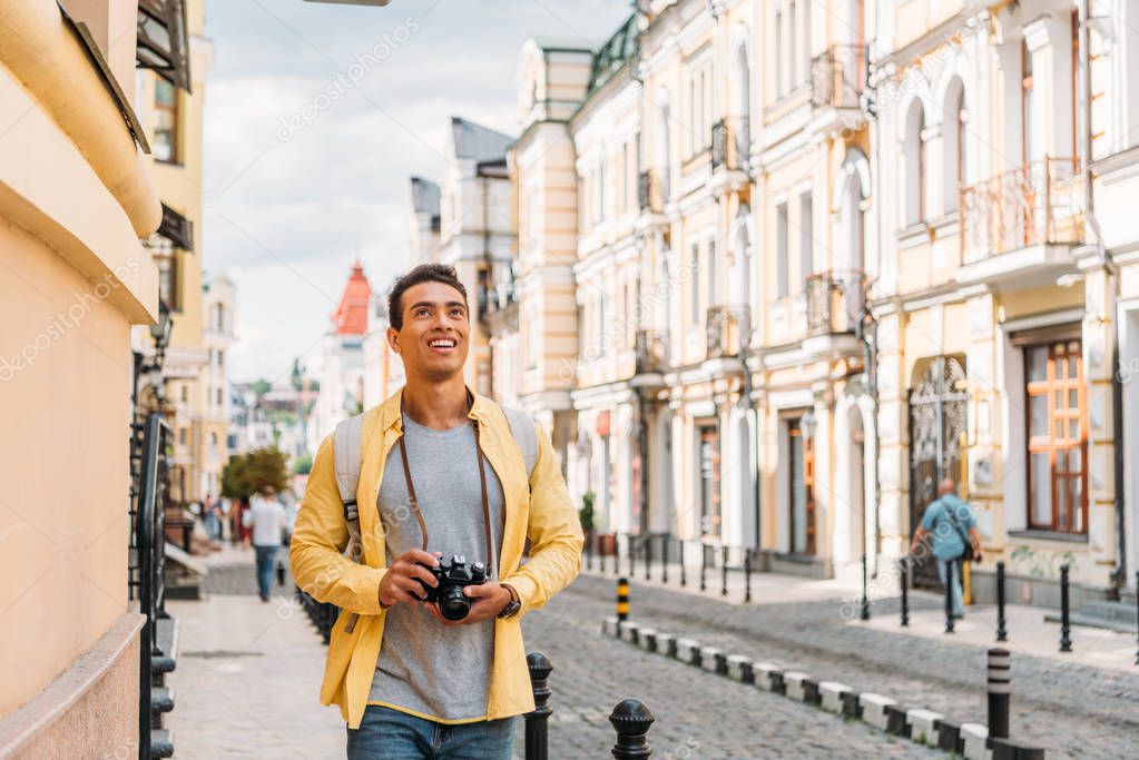 happy mixed race man smiling while holding digital camera near buildings 