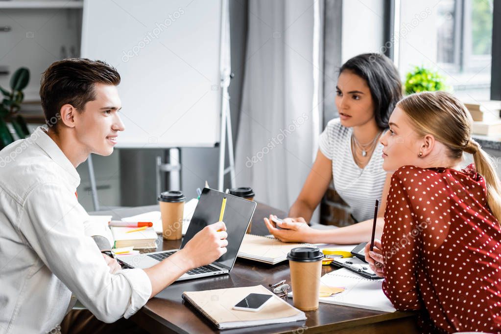 three multiethnic friends sitting at table and talking in apartment  