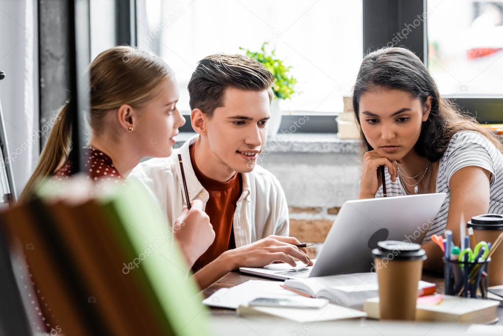 selective focus of multiethnic students smiling and looking at laptop in apartment 
