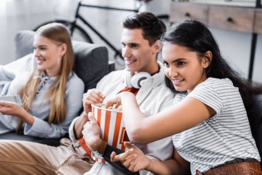 multicultural friends sitting on sofa and eating popcorn in apartment  clipart