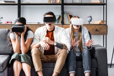 shocked multicultural friends with virtual reality headsets sitting on sofa 