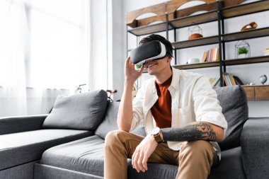 man with virtual reality headset sitting on sofa in apartment  clipart