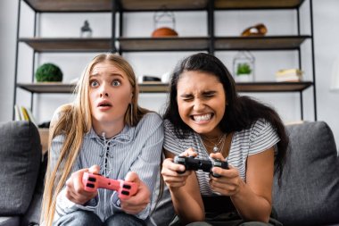 KYIV, UKRAINE - JULY 10, 2019: multicultural friends sitting on sofa and playing video game in apartment  clipart