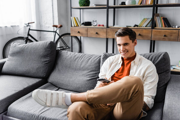handsome man in shirt sitting on sofa and holding remote controller in apartment 