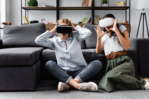 multicultural friends with virtual reality headsets sitting on floor in apartment 