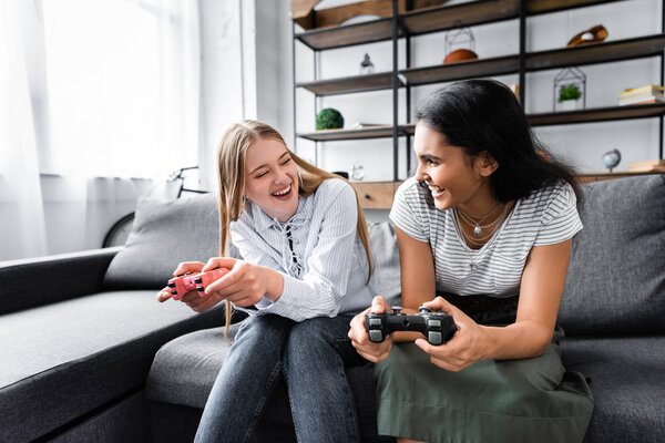 KYIV, UKRAINE - JULY 10, 2019: multicultural friends sitting on sofa and playing video game in apartment 