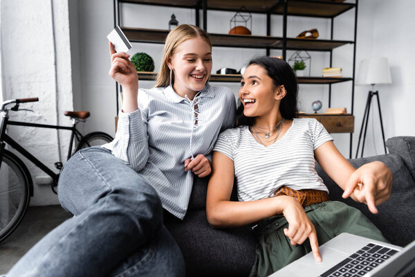 multicultural friends holding credit card and using laptop in apartment 