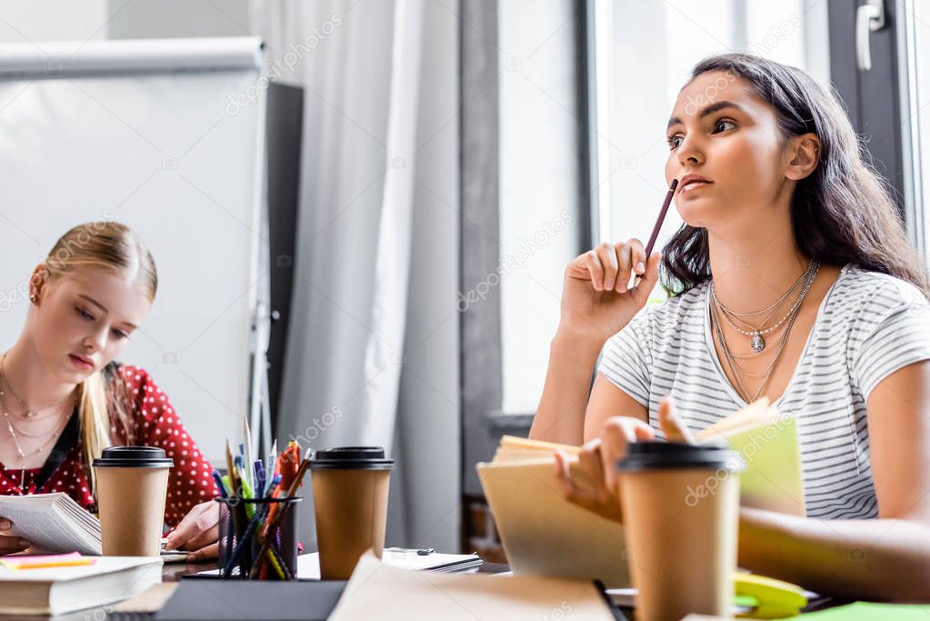 attractive multicultural friends sitting at table and studying in apartment 