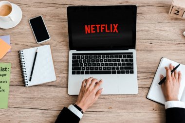 Kiev, Ukrayna - 23 Temmuz 2019: boş ekranlı akıllı telefonun yanındaki ekranda netflix web sitesi olan dizüstü bilgisayar kullanan kadının üst görünümü