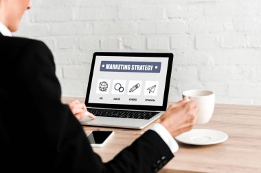 boş ekranlı pazarlama stratejisi yazıve akıllı telefon ile dizüstü bilgisayar yakınında otururken bardak tutan kadının seçici odak
