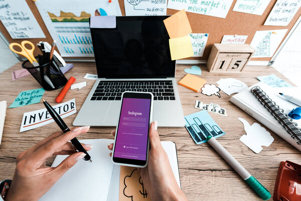 KYIV, UKRAINE - JULY 23, 2019: cropped view of woman holding smartphone with instagram app near sticky notes with lettering and laptop with blank screen 