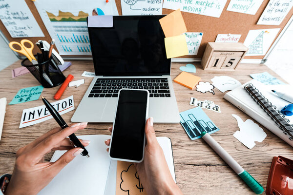 cropped view of businesswoman holding smartphone with blank screen near notebook and laptop with sticky notes in office 