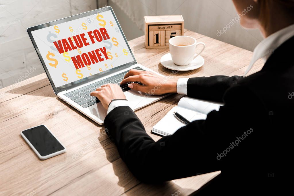 selective focus of woman typing on laptop with value for money letters near wooden cubes with calendar and smartphone with blank screen 