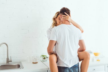 mutfak tezgahında oturan kız arkadaşı kucaklayan ve öpen genç adam geri görünümü