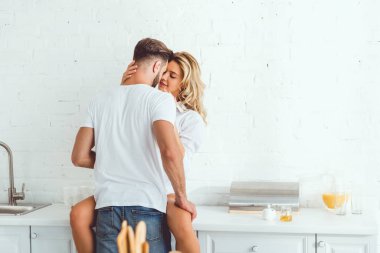 mutfak tezgahında oturan gülümseyen kız arkadaşı kucaklayan genç adamın arka görünümü