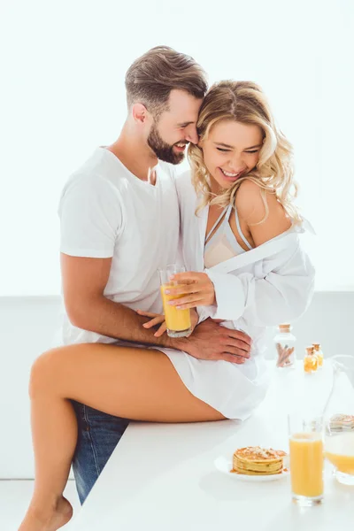 微笑的男人拥抱女友坐在厨房的桌子上 拿着一杯橙汁 — 图库照片