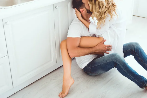cropped view of young couple hugging and kissing while sitting on floor in kitchen