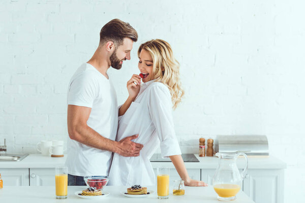 handsome young man embracing pretty girlfriend tasting strawberry in kitchen