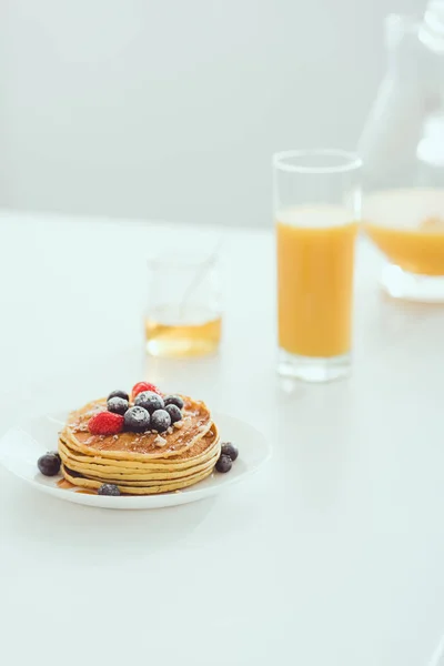 グラスの近くにおいしいパンケーキとベリーとプレートの選択的な焦点とオレンジジュースと白いテーブルに蜂蜜と瓶のジャグ — ストック写真