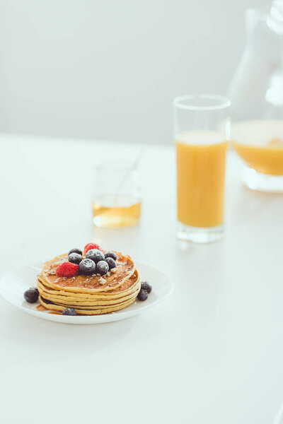 selective focus of plate with tasty pancakes and berries near glass and jug of orange juice and jar with honey on white table