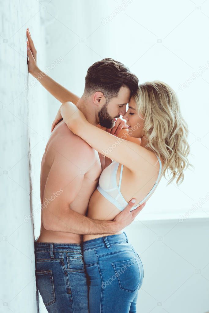 shirtless man and young woman in bra hugging while standing near white wall at home