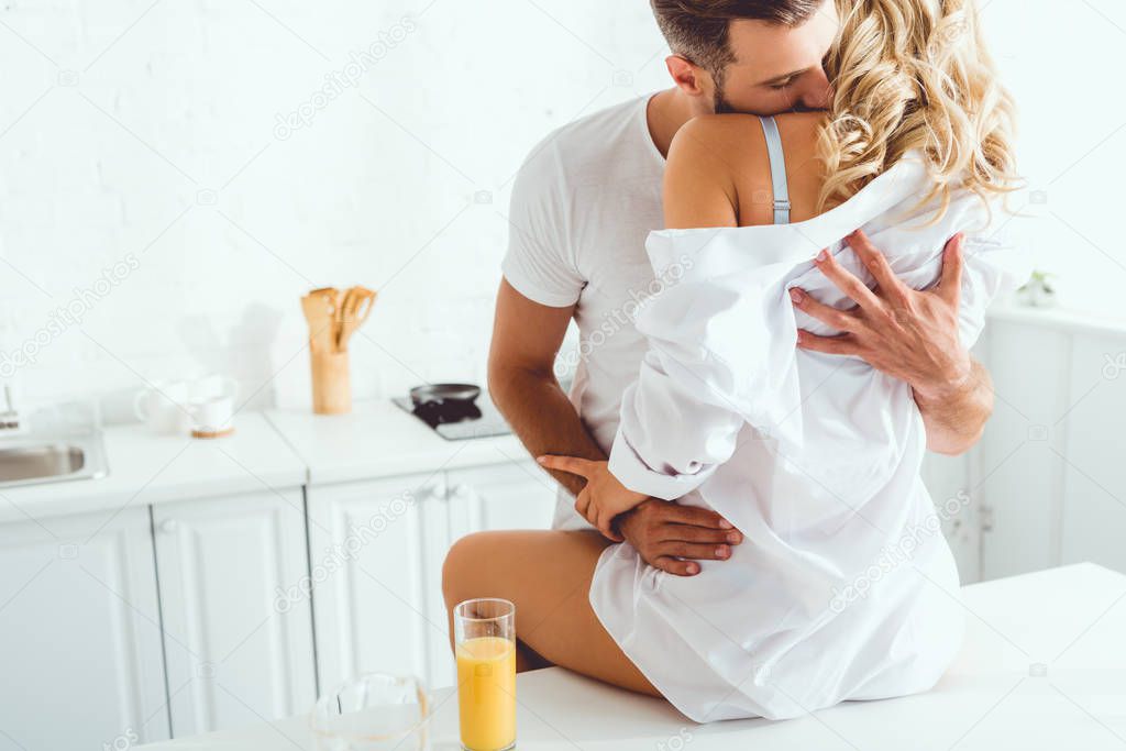 young man embracing and kissing girlfriend sitting on kitchen table near glass of orange juice