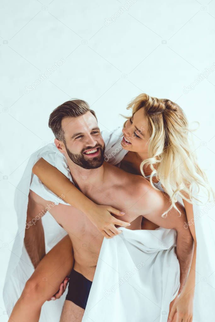 cheerful young man piggybacking smiling girlfriend while wrapping in white sheet