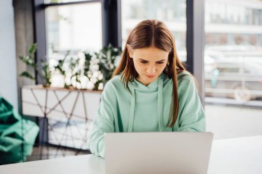 focused girl in casual hoodie sitting at table and using laptop clipart