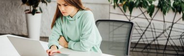 panoramic shot of upset girl sitting at table and using laptop at home clipart