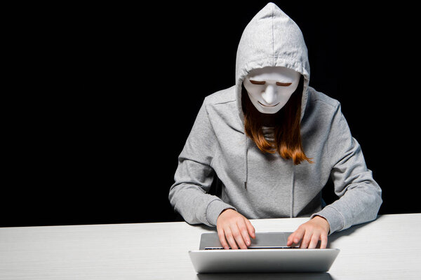 анонимный интернет-тролль в маске, печатающий на клавиатуре ноутбука изолированный на черном
