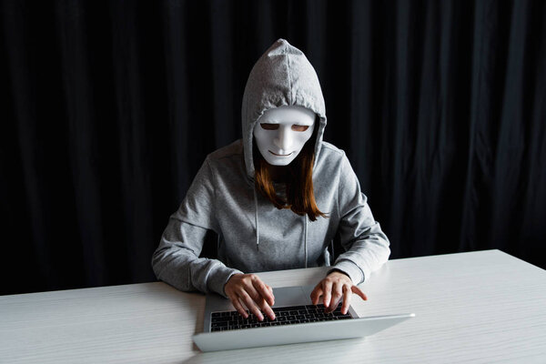 анонимный интернет-тролль в маске, печатающий на клавиатуре ноутбука на черном
