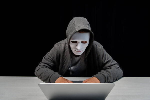 anonymous internet troll in mask typing on laptop keyboard isolated on black