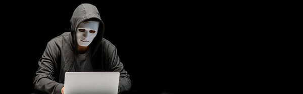 панорамный снимок анонимного интернет-тролля в маске, печатающего на клавиатуре ноутбука, изолированного на черном
