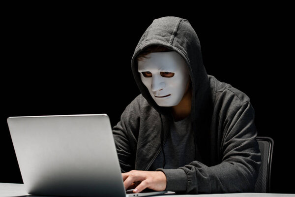 анонимный интернет-тролль в маске, печатающий на клавиатуре ноутбука изолированный на черном
