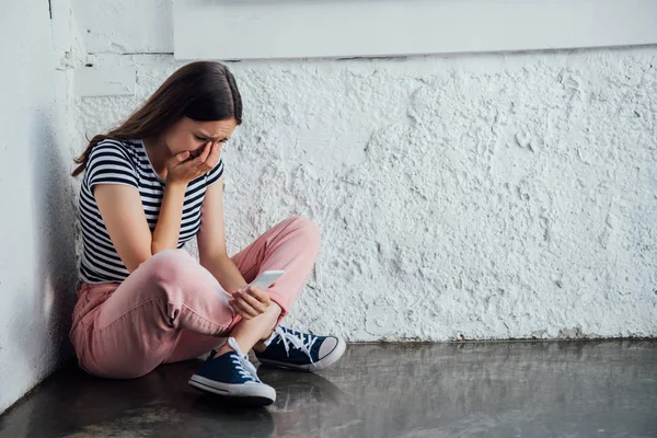 壁の近くに座って スマートフォンを保持しているピンクのズボンで悲しい泣く女の子 — ストック写真