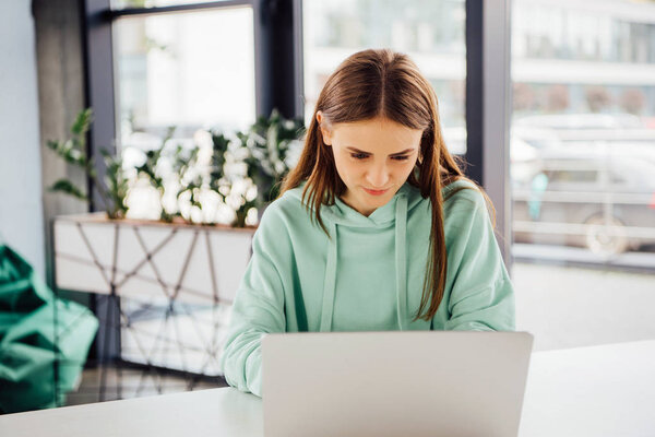 focused girl in casual hoodie sitting at table and using laptop