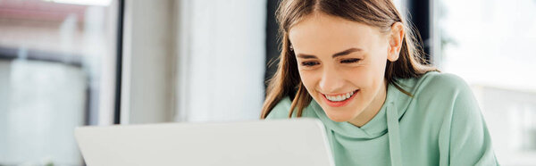 panoramic shot of smiling girl in casual hoodie sitting at table and using laptop