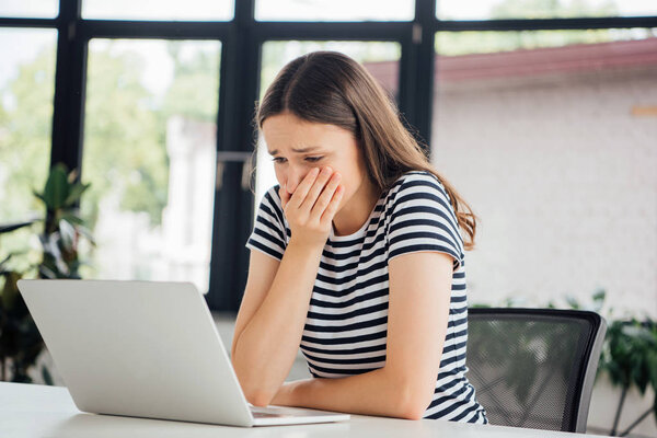 грустная девушка в полосатой футболке, прикрывающей рот рукой во время использования ноутбука дома
