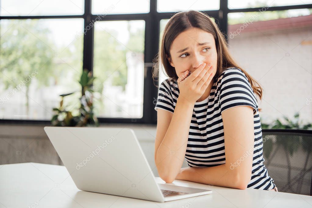 sad girl in striped t-shirt covering mouth with hand while using laptop at home