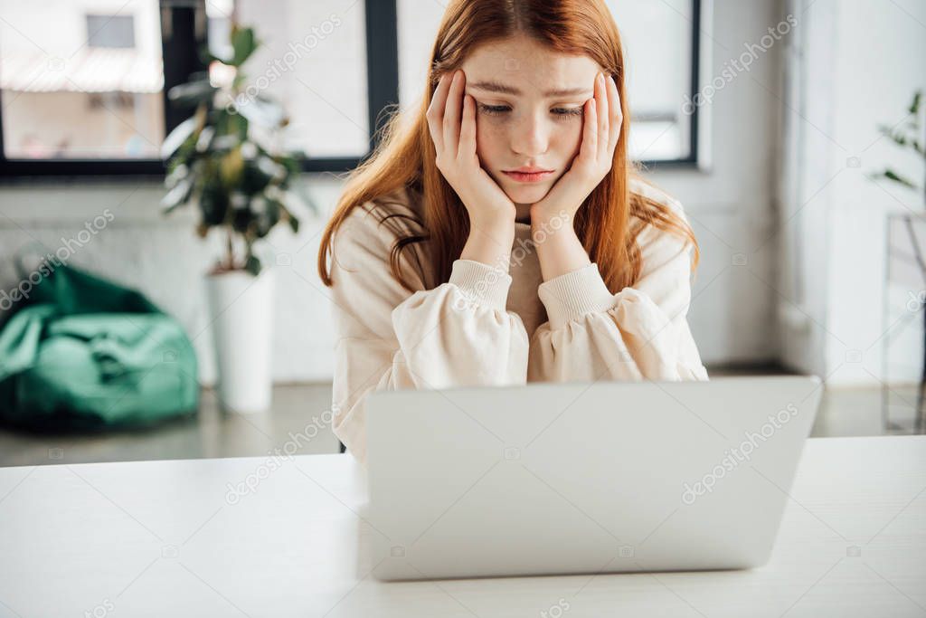 sad teen girl sitting at table with laptop at home