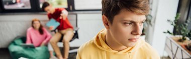 panoramic shot of pensive boy in yellow hoodie and laughing classmates bullying him clipart