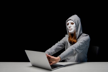 maske ve kapüşonlu anonim kız dizüstü bilgisayar yakınında oturan ve siyah izole siber zorbalık sırasında elleri germe