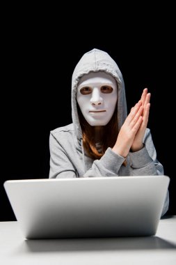 front view of anonymous girl in mask and hoodie sitting near laptop and rubbing hands during cyberbullying isolated on black clipart