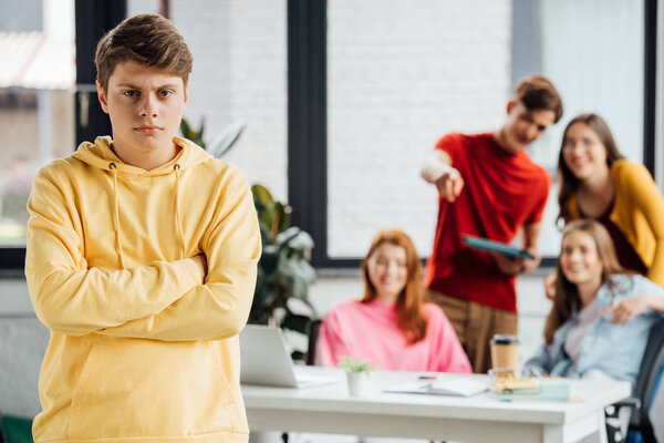 sad boy in yellow hoodie with crossed arms and laughing teenagers at desk