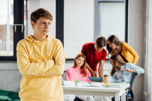 sad boy in yellow hoodie with crossed arms and laughing teenagers at desk