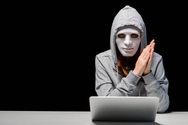 front view of anonymous girl in mask and hoodie sitting near laptop and rubbing hands during cyberbullying isolated on black