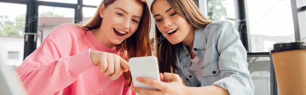 panoramic shot of two excited girls smiling while using smartphone in school